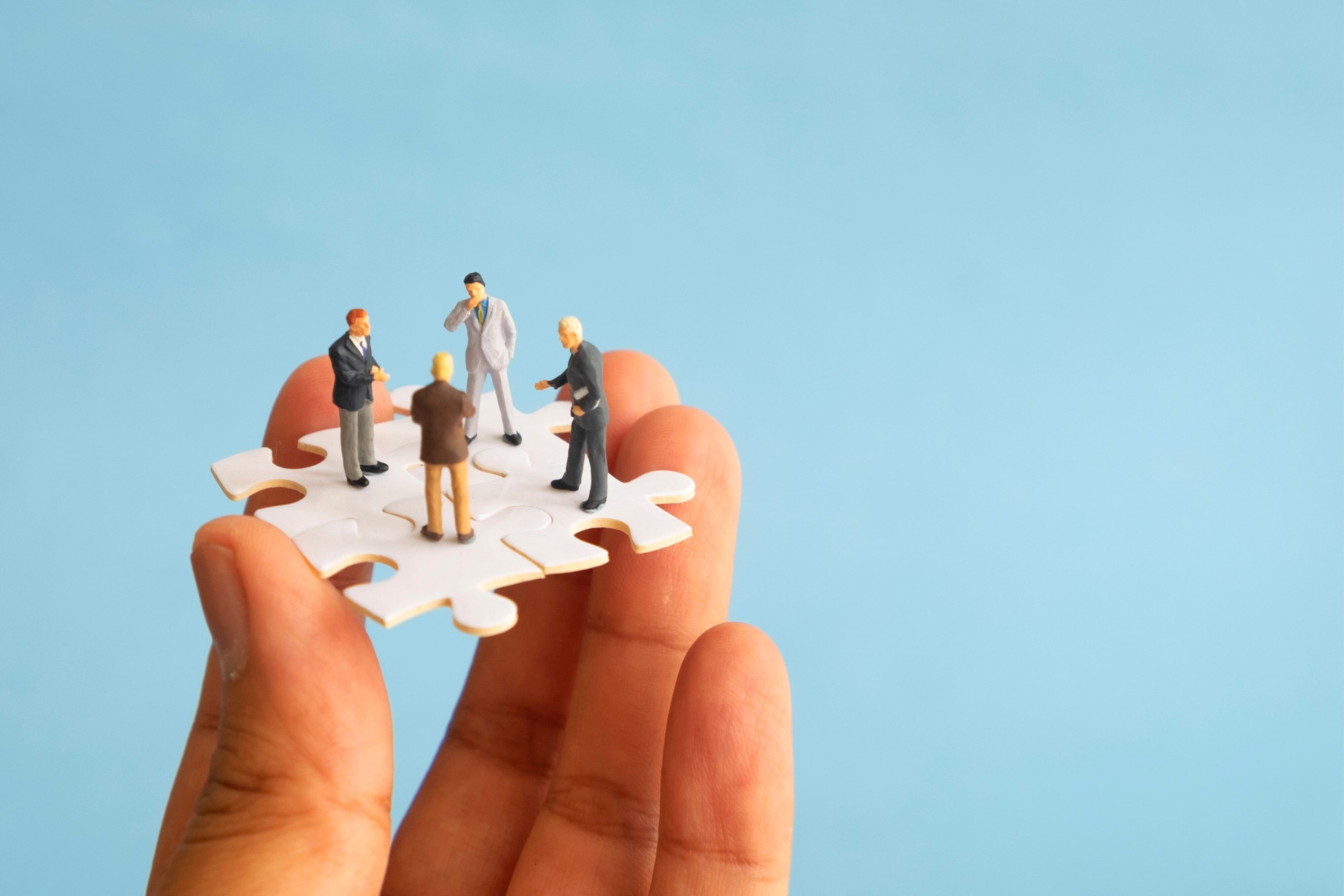 A hand holding a puzzle piece with miniature businesspeople standing on it, symbolizing teamwork, collaboration, and overcoming AI paralysis in the workplace.